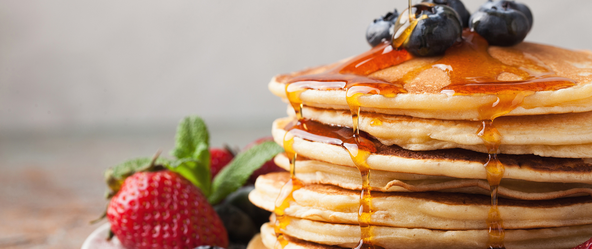 Close-up delicious pancakes, with fresh blueberries, strawberries and maple syrup on a light background. With copy space. Sweet maple syrup flows from a stack of pancake